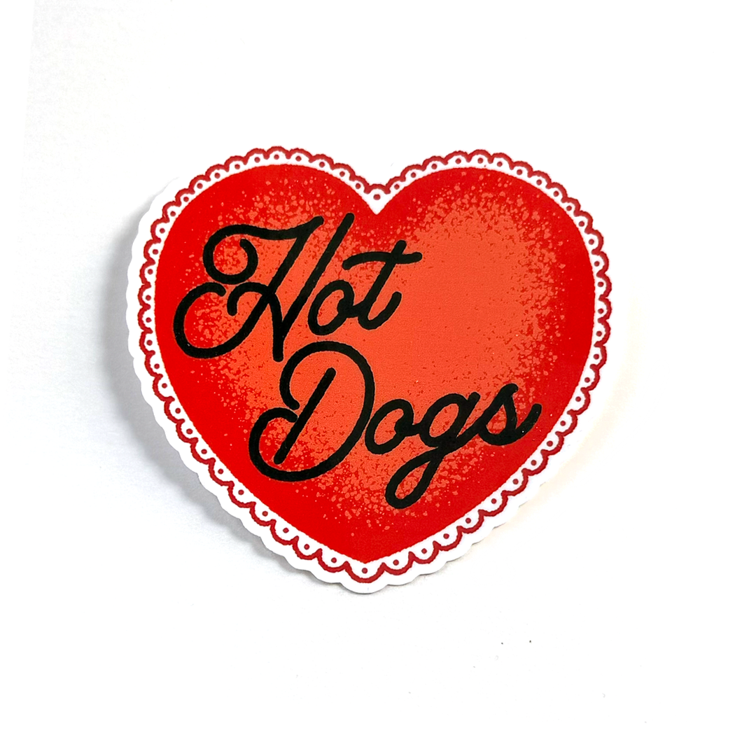 A heart-shaped sticker with a red center, black cursive text reading 'Hot Dogs', and a white scalloped border on a plain white background.